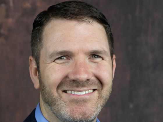 Man sites before a rust colored backdrop in a light blue shirt with navy pinstripe jacket