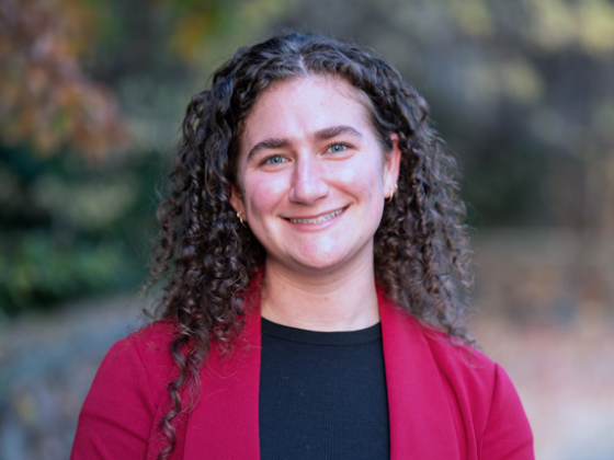 Woman with blue eyes and dark wavy hair stands outside in a burgundy jacket and black shirt.