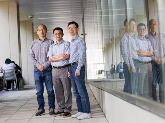 Three faculty members stand in an outdoor building corridor against a glass window with their reflections visible.