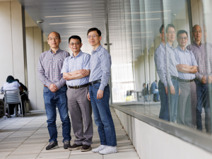 Three faculty members stand in an outdoor building corridor against a glass window with their reflections visible.