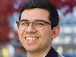 A man in grey glasses, blue checkered shirt, and blue blazer stands in an outdoor setting with red trees behind.